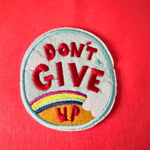 May include: A round, blue embroidered patch with a rainbow, clouds, and the text "DON'T GIVE UP" in red.