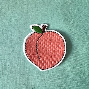 May include: Embroidered peach patch with a green leaf stem. The peach is a light pink colour and has a stitched outline.