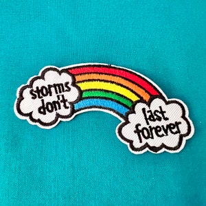 Op de afbeelding: Een geborduurde patch met een regenboog en witte wolken. De linker wolk zegt "storms don't" en de rechter wolk zegt "last forever".