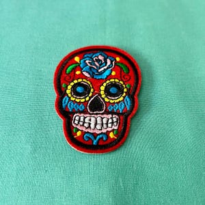 May include: Red embroidered sugar skull patch with blue, yellow, and green details. The skull has a flower on the forehead and a toothy grin.