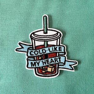 May include: An embroidered patch featuring a cartoon illustration of a cold drink with a straw and ice cubes. The drink is surrounded by a blue ribbon with the text "Cold Like My Heart".