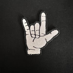 May include: White embroidered patch of a hand making the 'I love you' sign language gesture.