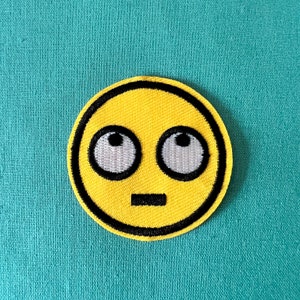 May include: Yellow embroidered iron-on patch with a cartoon face with wide eyes and a straight mouth. The face has a black outline.