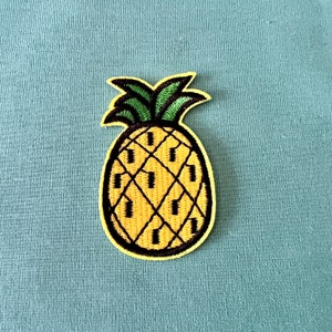 Op de afbeelding: Een gele ananas met een groene, bladerrijke bovenkant, geborduurd op een blauwe stoffen achtergrond.