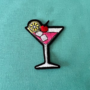 Peut inclure: Patch brodé thermocollant représentant un cocktail rose avec un quartier de citron et une cerise sur le dessus.