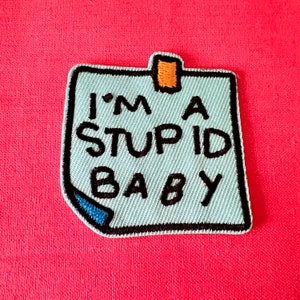 May include: A light blue patch in the shape of a sticky note with the text "I'M A STUPID BABY" in black. The patch has a black border, a blue corner, and an orange tab. The background is a solid red.