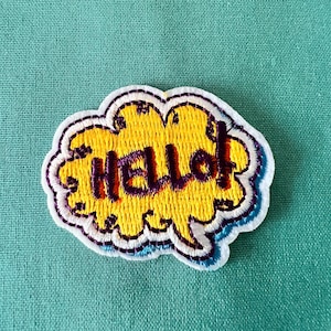 Op de afbeelding: Een gele geborduurde patch in de vorm van een spraakballon met het woord "Hello!" in paarse draad.