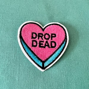 May include: A pink heart-shaped patch with blue trim and the text "DROP DEAD" in black embroidery.