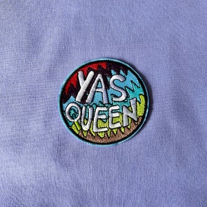 May include: A round, colourful embroidered patch with the text "YAS QUEEN" in white letters. The patch has a blue background with green, yellow, orange, and purple accents.