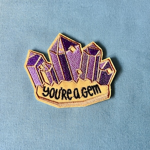Op de afbeelding: Paarse en gouden geborduurde strijkpatch met een cluster kristallen en de tekst "You're a Gem".