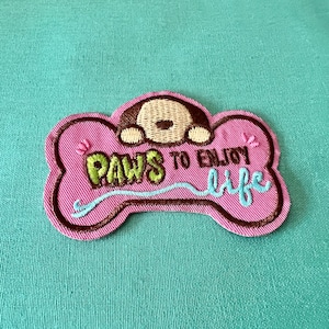 May include: A pink embroidered patch in the shape of a dog bone. The patch features a brown dog face, the words "PAWS TO ENJOY life" in green and blue, and a brown border.