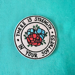 May include: White embroidered patch with a red and blue flower design. The text around the flower reads "There is strength in your softness."