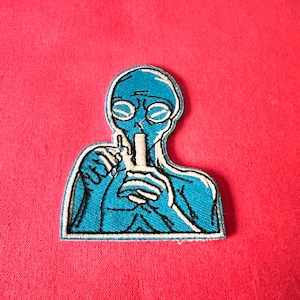 Könnte beinhalten: Blauer gestickter Aufnäher eines Außerirdischen mit Brille, der eine Glaspfeife hält.
