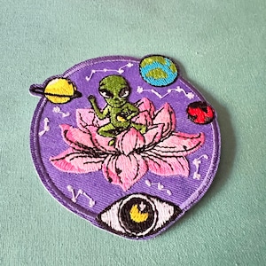 Könnte beinhalten: Lilafarbener gestickter Aufnäher mit einem grünen Außerirdischen, der auf einer rosa Lotusblüte sitzt. Der Außerirdische hält eine kleine gelbe Kugel. Im Hintergrund befinden sich drei Planeten und Sterne. Ein weißes Auge mit schwarzer Pupille und einem gelben Halbmond befindet sich unten.