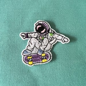 Può includere: Una toppa ricamata di un astronauta che indossa una tuta spaziale bianca e un casco, mentre fa skateboard su uno skateboard viola e giallo.