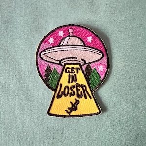 May include: An embroidered patch featuring a UFO and the text "Get in Loser" in pink and yellow.