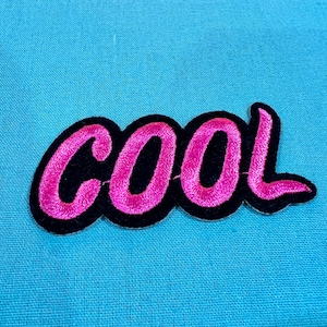 Pode incluir: Etiqueta bordada rosa e preta com a palavra "COOL" em uma fonte estilizada.