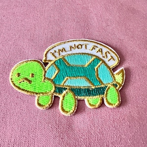 Może przedstawiać: Naszywka z haftem przedstawiająca zieloną i niebieską żółwia z napisem "I'm not fast" na białym banerze nad żółwiem.
