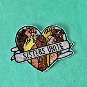 May include: Embroidered patch in the shape of a heart with the text "Sisters Unite" in a banner. The heart features five hands of different skin tones clasped together.