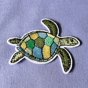 May include: An embroidered sea turtle patch in green, blue and yellow. The turtle is outlined in black thread.