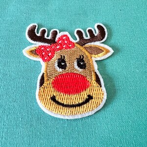 May include: Embroidered iron-on patch of a reindeer with a red and white polka dot bow. The reindeer has brown antlers, a red nose, and a brown face with black eyes and a smile.