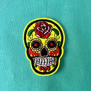 May include: Yellow embroidered sugar skull patch with a red rose on the forehead, black eyes, and white teeth. The skull is decorated with colorful details.