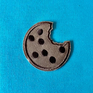 May include: A brown fabric cookie with a bite taken out of it. The cookie has six black chocolate chips.