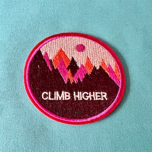 May include: A pink and red embroidered patch with a mountain range and a sun. The text "CLIMB HIGHER" is embroidered in white on the patch.