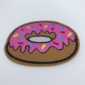 May include: A pink and brown embroidered donut patch with colorful sprinkles. The donut has white frosting and a hole in the center.