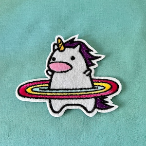 May include: White unicorn with a purple mane and a yellow horn, standing inside a rainbow-colored ring. The unicorn is wearing a pink nose and has a black outline.