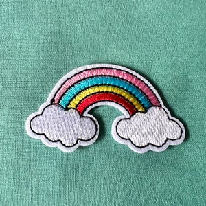 May include: A white felt rainbow patch with pink, blue, yellow, red, and green embroidery. The rainbow has white clouds on either side.