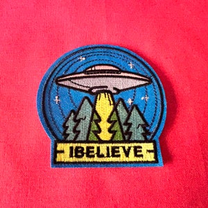 May include: A blue and green embroidered patch featuring a UFO flying over a forest with the text "I BELIEVE" in yellow.