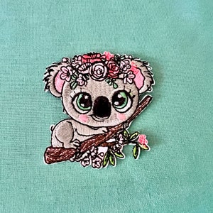 May include: Embroidered iron-on patch of a koala bear wearing a pink flower crown and sitting on a branch with pink flowers.