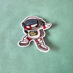 May include: Embroidered iron-on patch of an astronaut in a white spacesuit with a red stripe across the chest, a yellow patch on the chest, and a black helmet. The astronaut is in a dancing pose with one arm raised and the other arm extended to the side.
