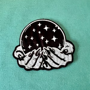 May include: Embroidered patch depicting a black sphere with white stars, held by two white hands. The patch has a black border and is set against a light teal background. The design has a mystical theme.