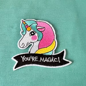 Pode incluir: Etiqueta bordada para passar a ferro com um unicórnio de desenho animado com uma crina rosa e azul e um chifre amarelo. O unicórnio está segurando uma faixa que diz "You're Magic!"