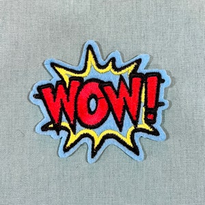 Peut inclure: Un patch brodé avec le mot "WOW!" dans une police de style bande dessinée, en rouge et jaune.