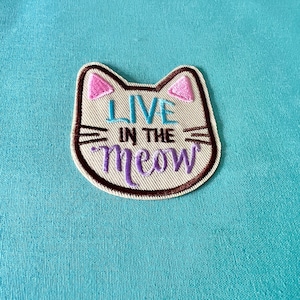 May include: A white cat-shaped patch with brown stitching and pink ears. The patch says "Live in the meow" in purple and blue embroidery.