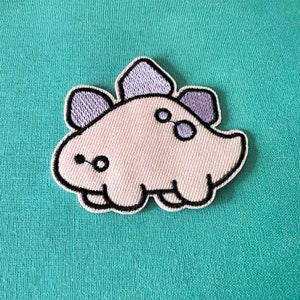 May include: A pink embroidered dinosaur patch with purple accents. The dinosaur has a cute cartoon-like expression with black eyes and a small mouth.