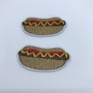 Op de afbeelding: Twee geborduurde strijkpatches van hotdogs met mosterd en ketchup. De patches zijn bruin, geel en rood.