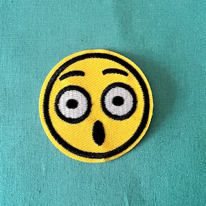 May include: Yellow embroidered patch with a surprised face with wide eyes and an open mouth. The face is outlined in black.