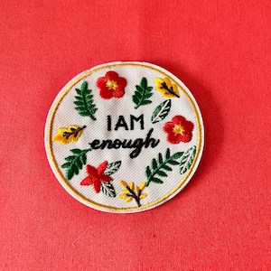 Op de afbeelding: Witte geborduurde patch met een bloemenmotief en de tekst "I AM enough". De bloemen zijn rood, geel en groen.