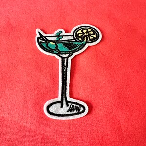 May include: A white felt embroidered patch of a green cocktail with a lemon wedge. The cocktail is in a martini glass with a black outline.