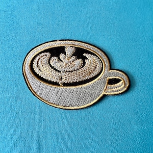 Può includere: Una toppa ricamata in oro e nero di una tazza di caffè con latte art. La tazza è bianca con un bordo e un manico neri. La latte art è un design a spirale dorato.