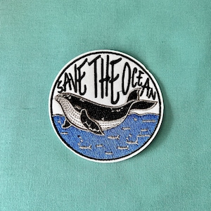 May include: A white embroidered patch featuring a black whale swimming in blue water. The text "Save The Ocean" is written in black around the whale.