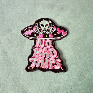 May include: Embroidered iron-on patch featuring a cartoon alien in a flying saucer with the text "NO BAD TRIPS" in pink and white lettering.