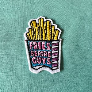 Può includere: Una toppa ricamata da stirare con un'immagine di cartone animato di un contenitore di patatine fritte con il testo "Fries Before Guys" in lettere rosa, blu e gialle.
