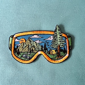 May include: Embroidered patch of a ski goggle with a mountain scene inside. The goggle is orange and yellow with a blue sky, green mountains, and a green pine tree on the right side.