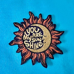 May include: A pink and purple embroidered patch with a sun and moon design. The text "You Are My Sunshine" is written in a white script font.