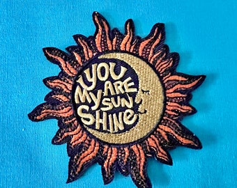 May include: A pink and purple embroidered patch with a sun and moon design. The text "You Are My Sunshine" is written in a white script font.